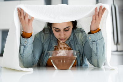 woman inhaling steam over bowl of hot water with white towel over her head