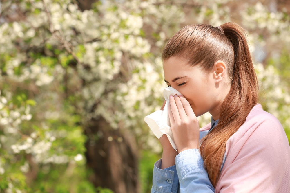 young woman with seasonal allergies sneezing into a tissue next to a blooming tree